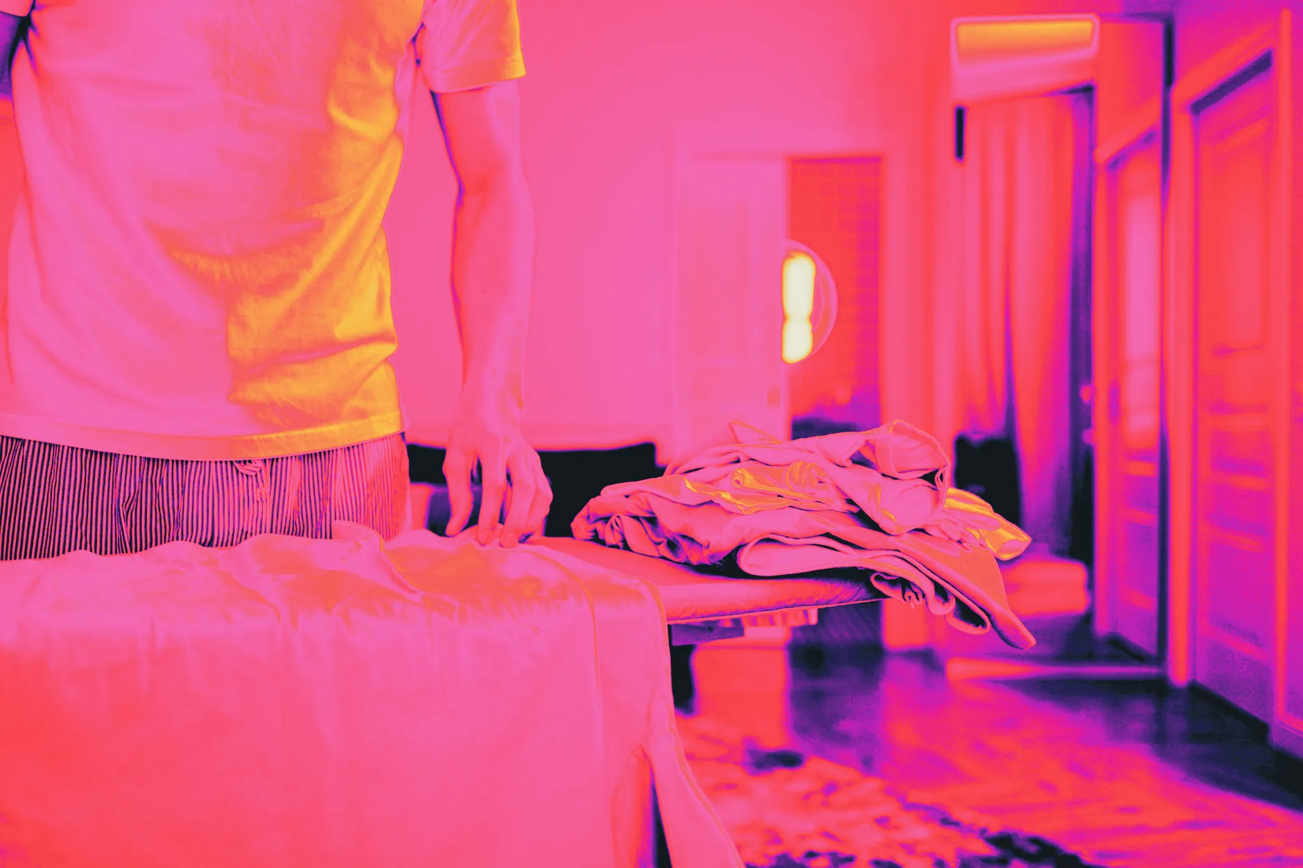 A person presses a garment on an ironing board, with a stack of folded laundry beside them